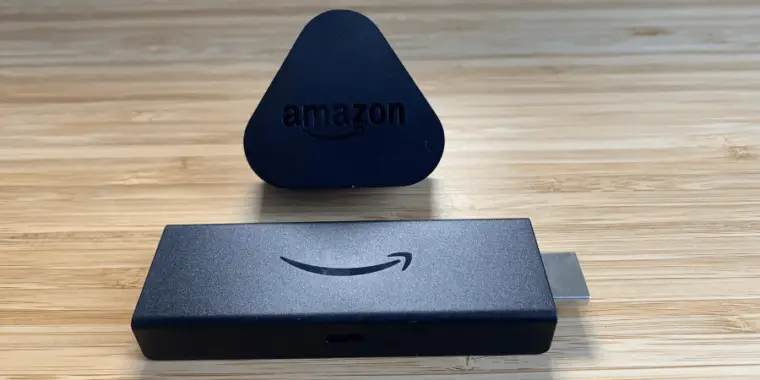 In the foreground is an unplugged black Firestick device with a HDMI connector on one side and the Amazon smile logo in the center. Behind it stands a small, black, triangular stand with the word "amazon" in white letters.