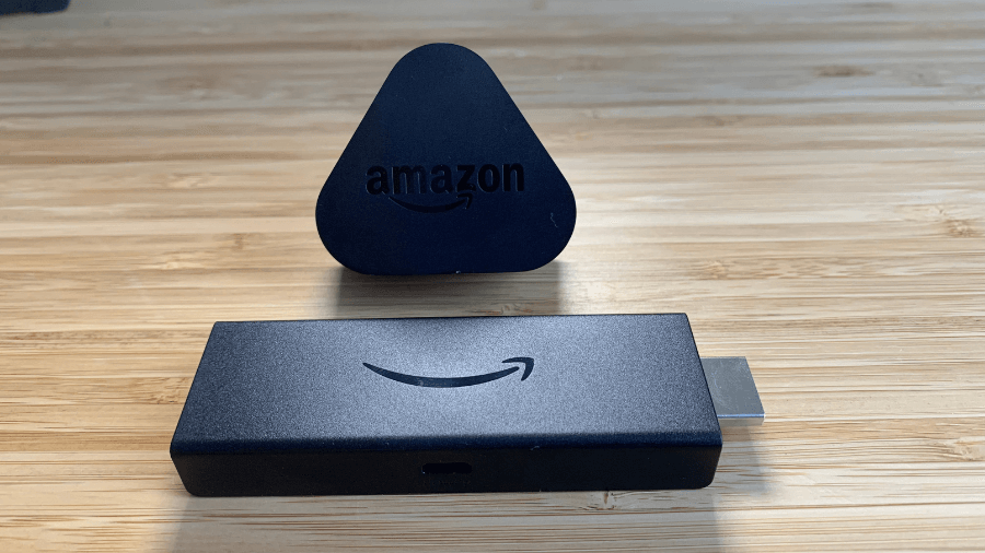 In the foreground is an unplugged black Firestick device with a HDMI connector on one side and the Amazon smile logo in the center. Behind it stands a small, black, triangular stand with the word "amazon" in white letters.