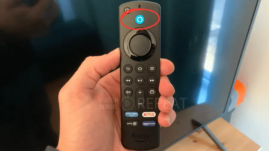 A person's hand holding a Fire TV remote with a blue Alexa ring illuminated at the top, pointing towards TV screen.