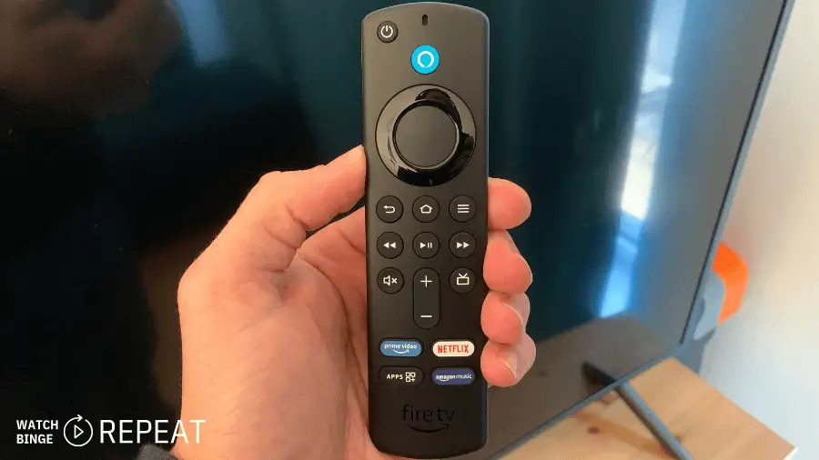 This image shows a person's hand holding a Fire TV remote control. The remote is black with a navigation circle at the top, a blue microphone button indicating voice control capability, and dedicated buttons for services like Amazon Prime Video and Netflix. There are also playback control buttons and a power button at the top. The TV in the background is reflecting a window, and there's a logo that says "WATCH BINGE REPEAT" in the corner of the image.