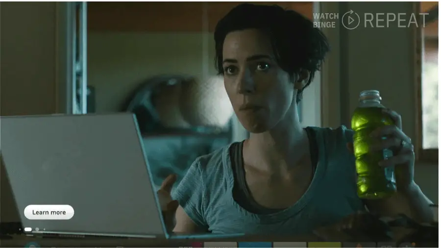 A woman sits at a table with a laptop, holding a bright green drink, with a thoughtful expression on her face.