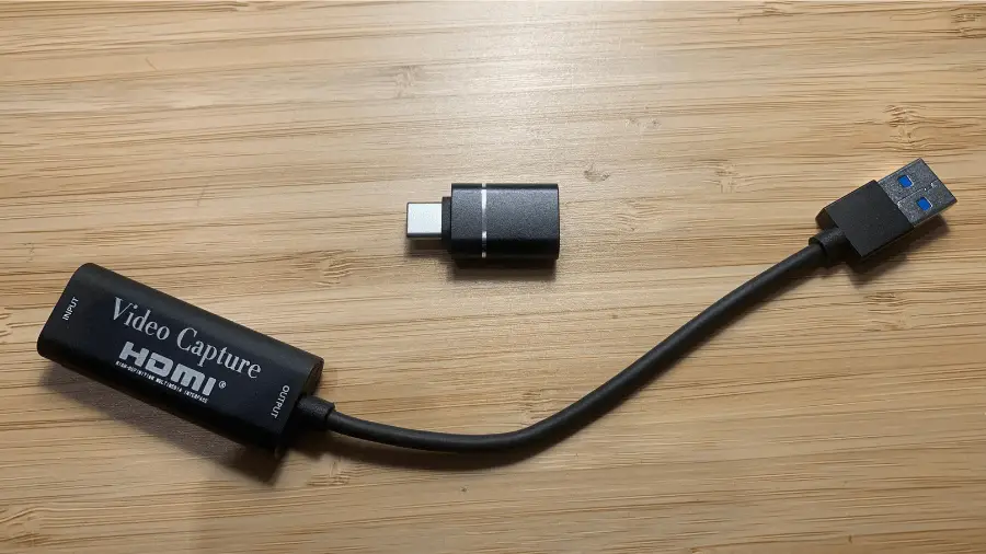 A USB video capture card with HDMI and USB-A connectors is shown against a wooden background.