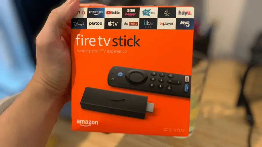 A person's hand holding a packaged Amazon Fire TV Stick. The box is orange with images of the device and remote, and logos for various streaming services.